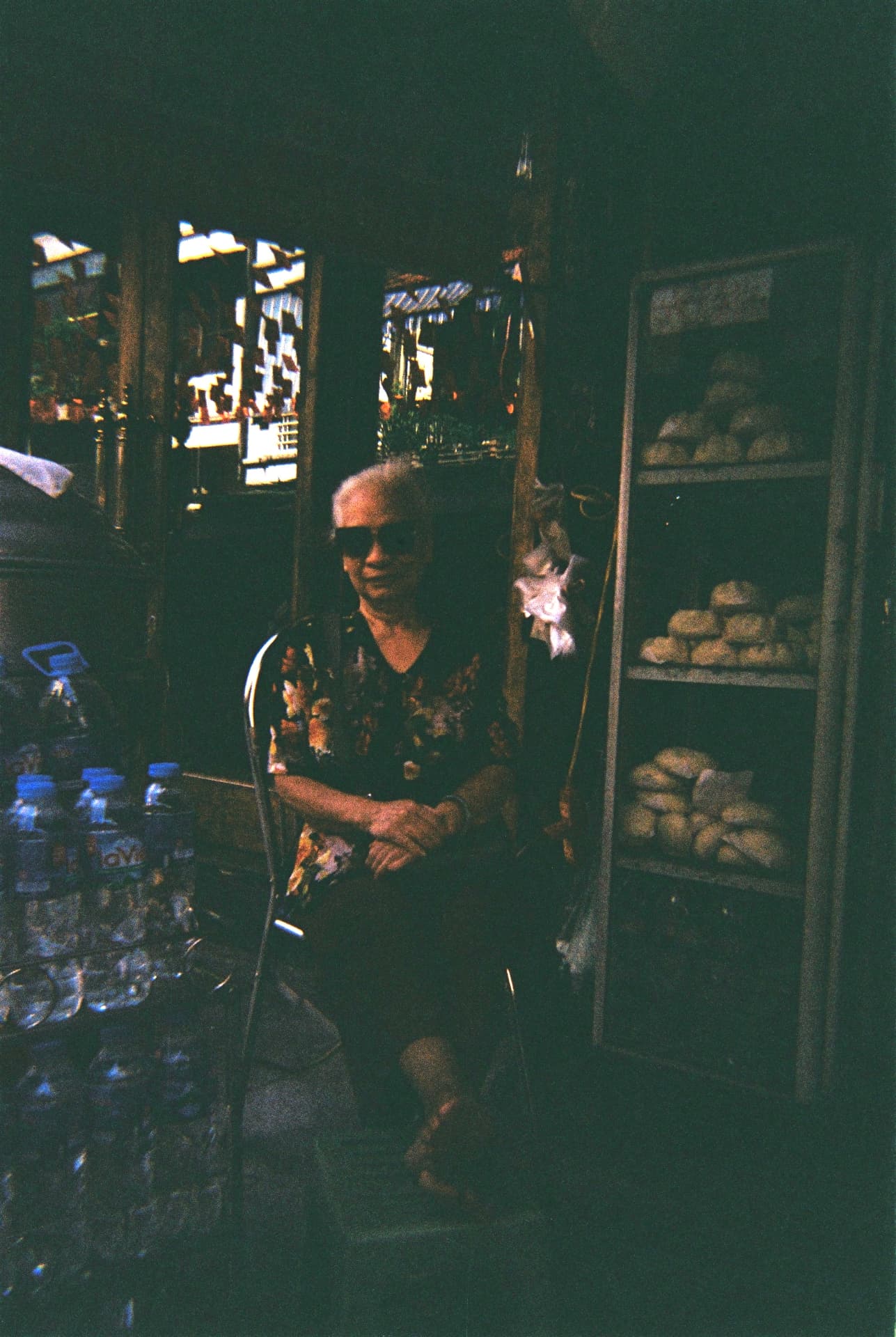 An older woman sitting in the shade of a dim market stall beside stacked water bottles and bread