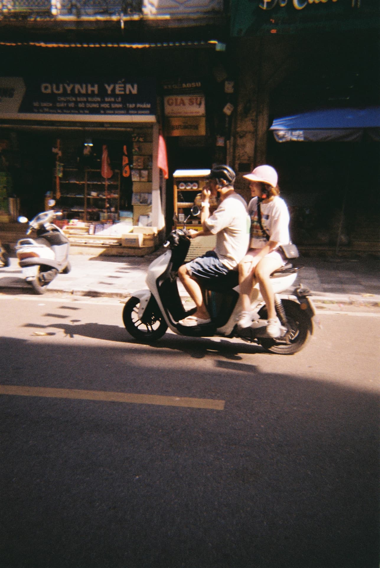 Two people riding a scooter past a shopfront in bright afternoon sun