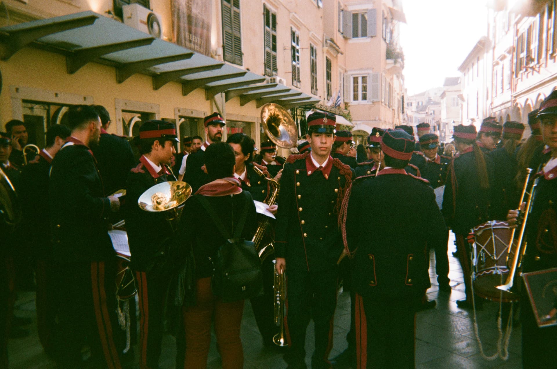 A marching band in dark uniforms gathering in a narrow sunlit street