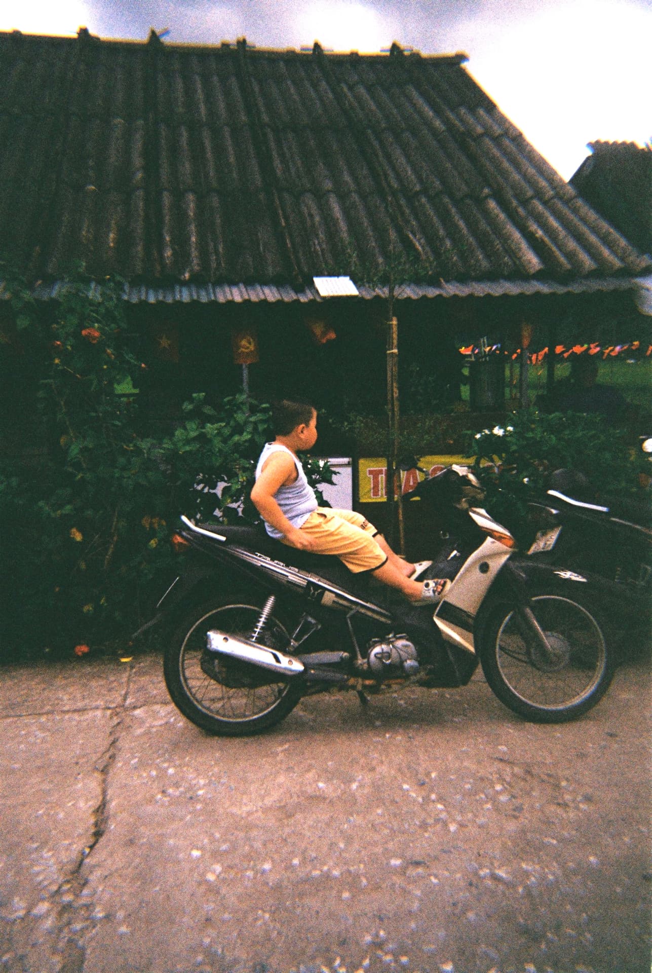 A boy sitting on a parked motorbike outside a tiled-roof house at dusk